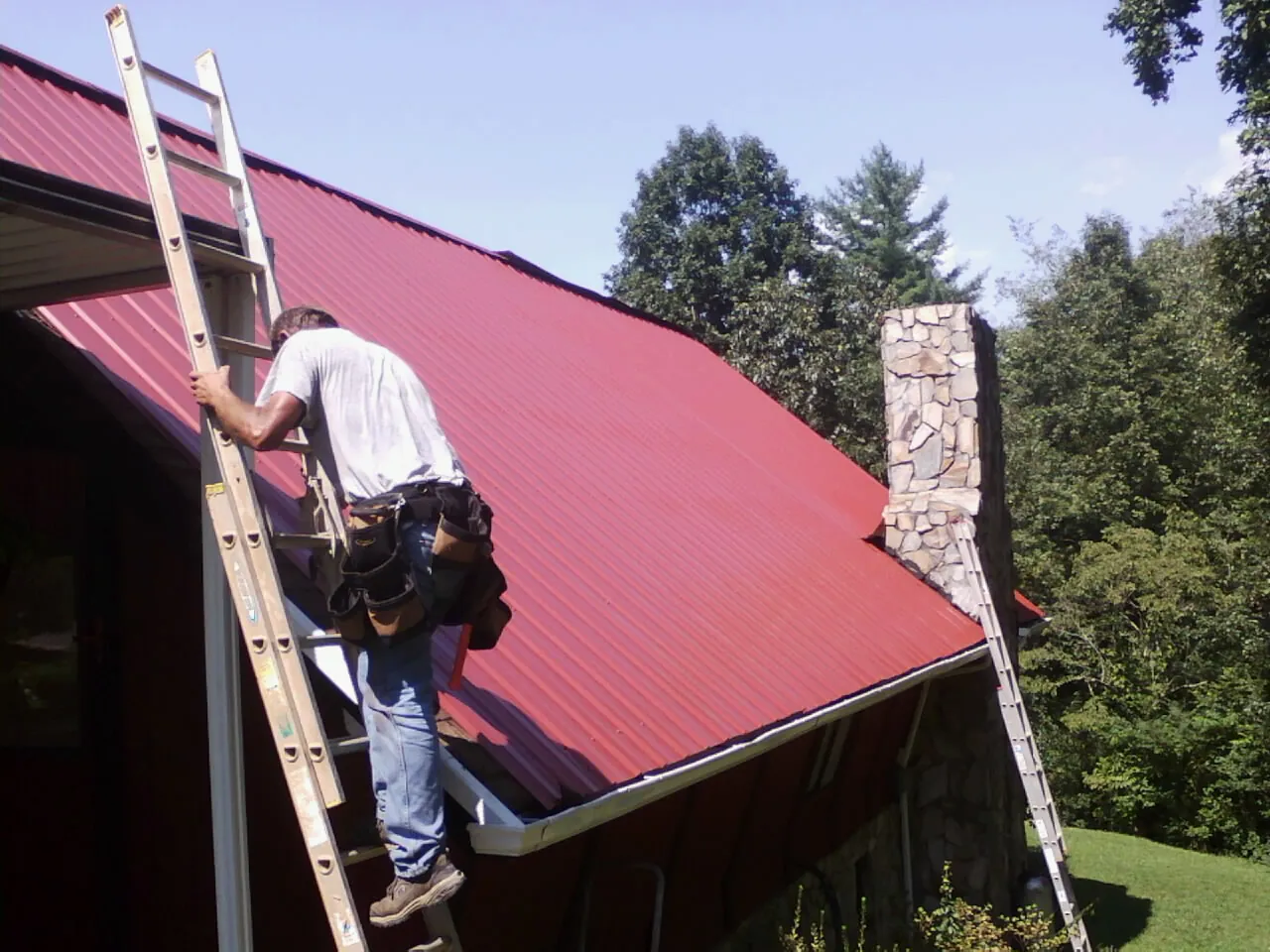 Roofer on a ladder beside a red metal roof and stone chimney — Soffit & Fascia Repair by Roofing Brothers in Perry Point
